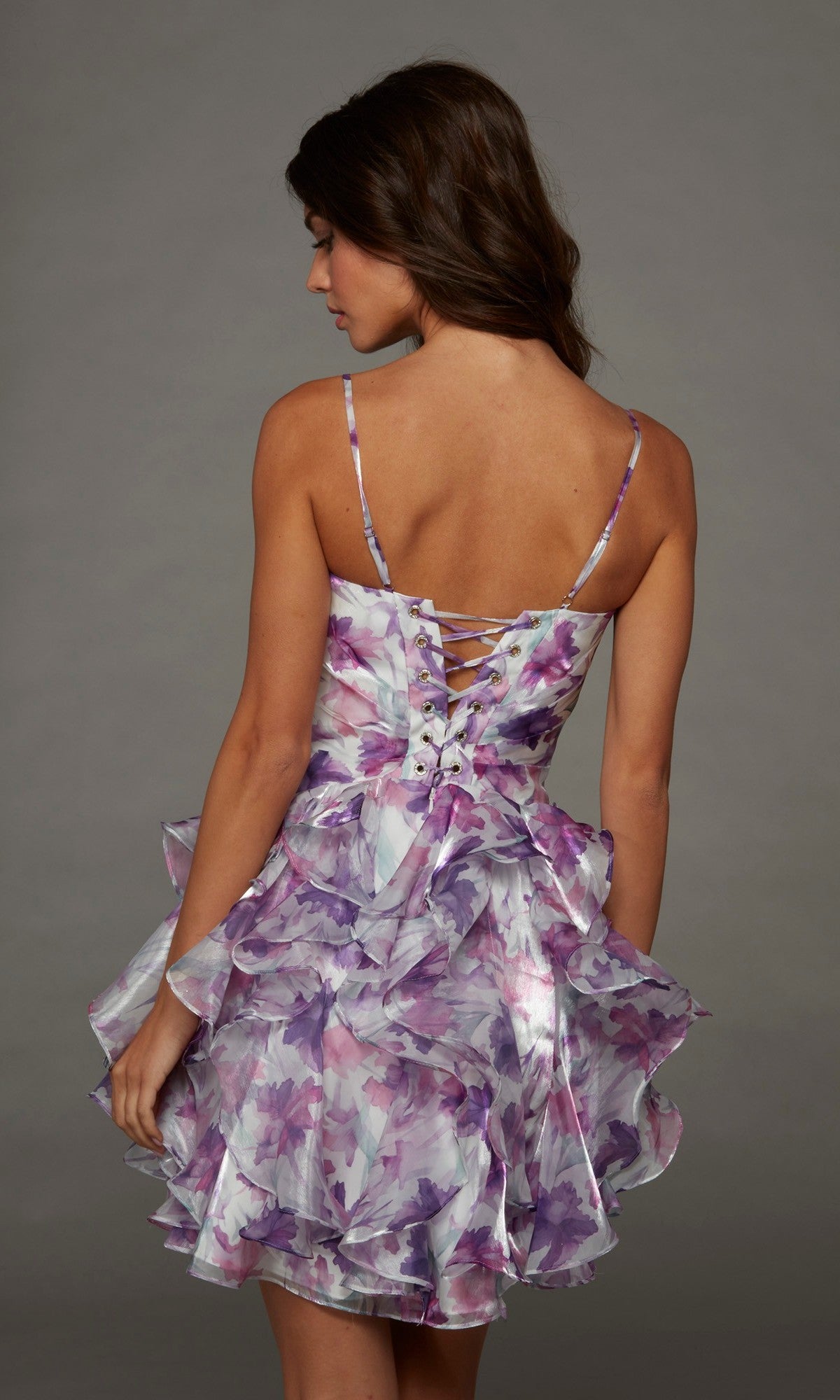 A woman stands with her back to the camera against a plain gray background, wearing the Alyce 30116 short homecoming dress—a sleeveless purple organza design featuring a lace-up back and a ruffled skirt.